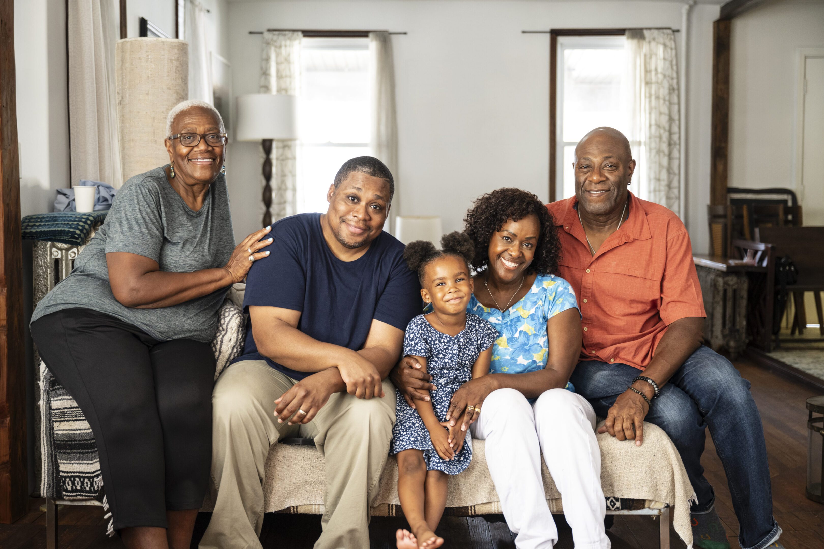 Front view of senior couple in their 60s, mature couple in 40s and 50s, 4 year old child, all together on sofa, looking at camera and smiling.
